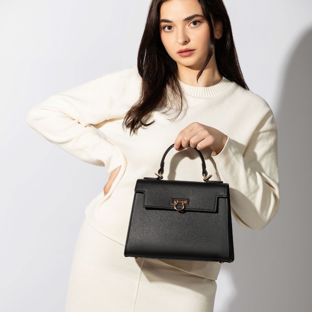 A woman in a white dress elegantly holds a black handbag from the Levantine top handle bags, Layla collection