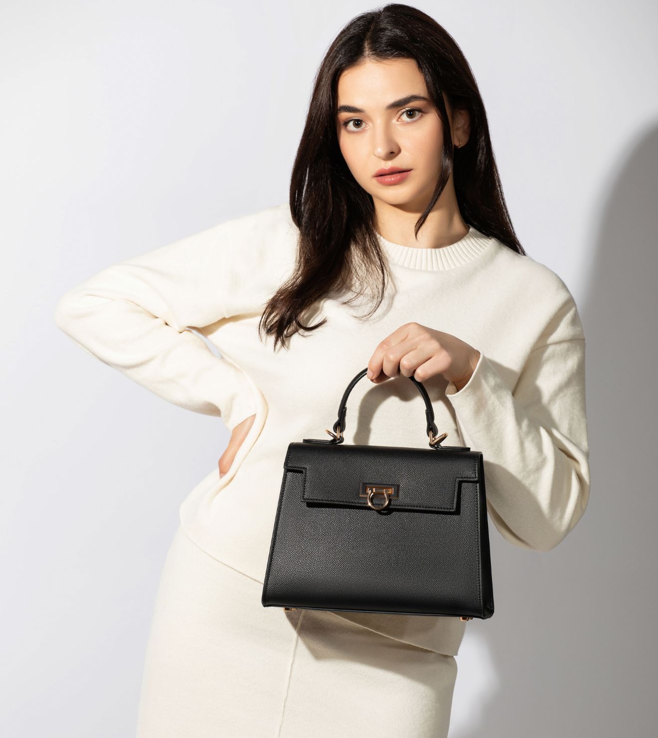 A woman in a white dress elegantly holds a black handbag from the Levantine top handle bags, Layla collection