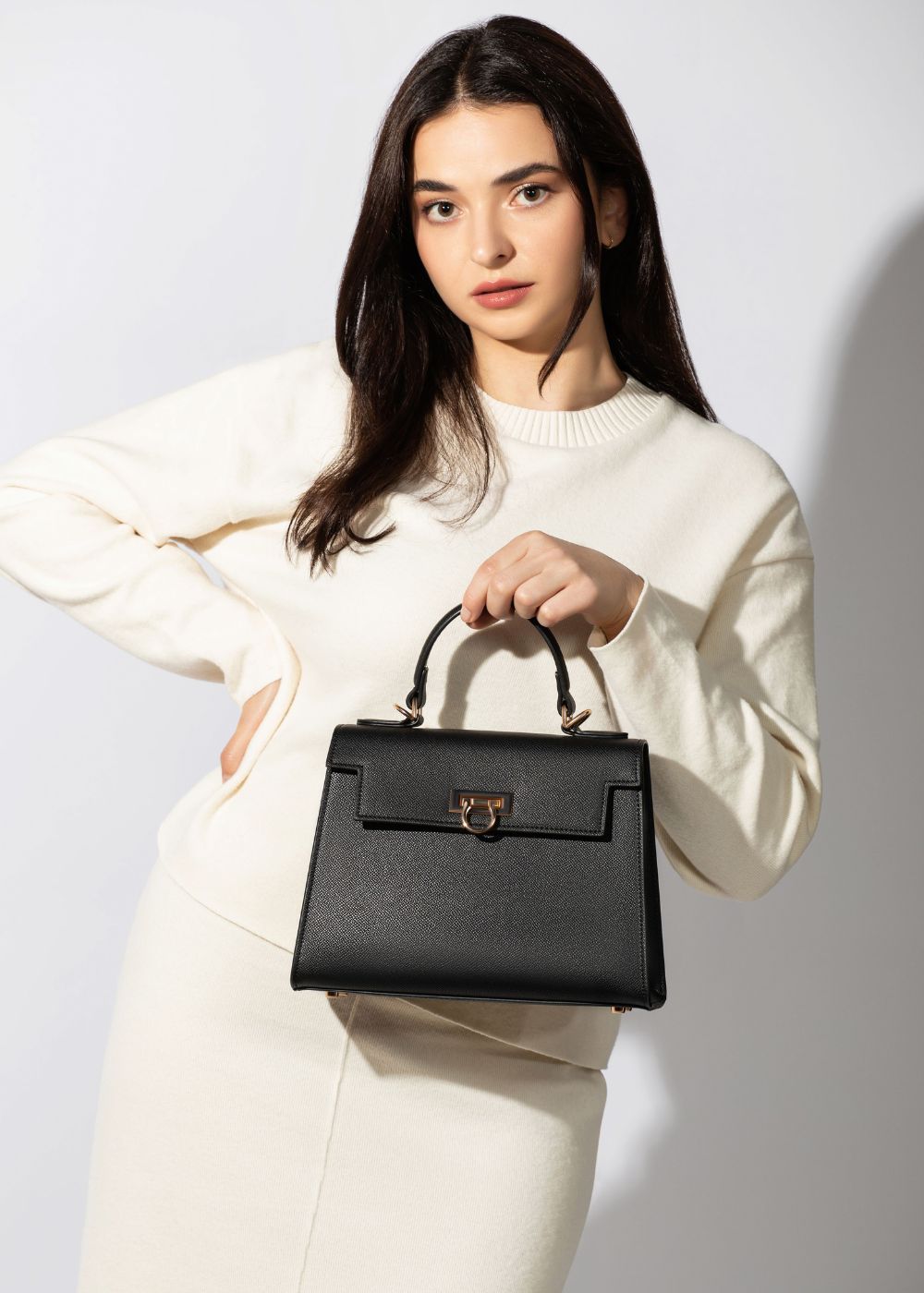 A woman in a white dress elegantly holds a black handbag from the Levantine top handle bags, Layla collection